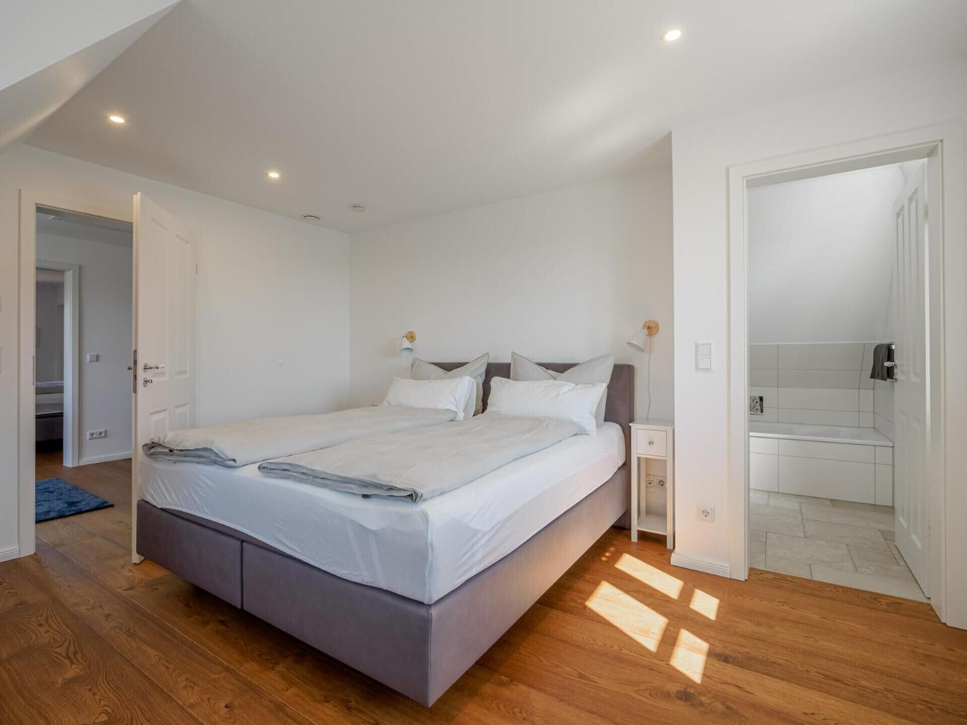 A bedroom with a bed, bedside tables, a nightstand, a bathroom with a shower, and a door leading to another room.