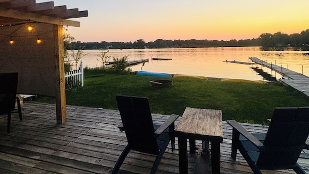 A wooden deck with two chairs and a table, overlooking a calm lake with a dock and a grassy area.