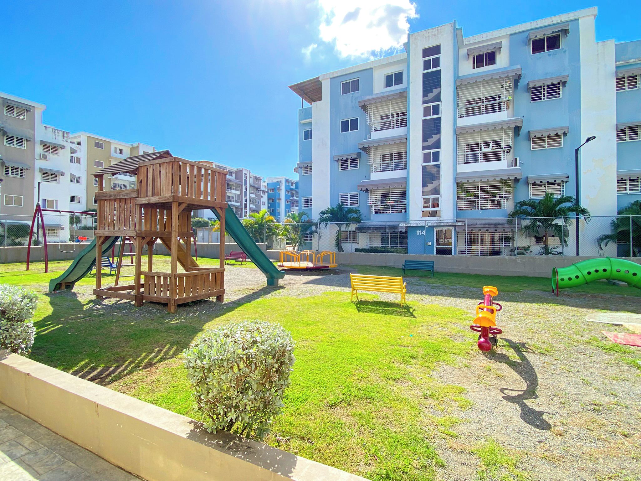 A playground with a wooden play structure, a green slide, and a yellow bench in front of a multi-story residential building.