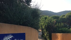 Placa de 'Les Palmier de Palombaggia Residence', com um caminho de cascalho serpenteando por uma área arborizada, com árvores e colinas ao fundo.