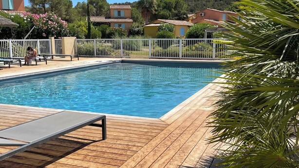 Uma piscina com deck de madeira, cercada por um muro branco e palmeiras.