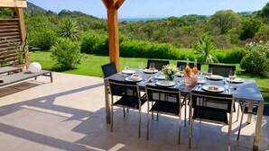 Uma área de refeições ao ar livre com uma mesa posta para dois, rodeada por cadeiras, com vista para uma paisagem exuberante e uma pérgola de madeira.
