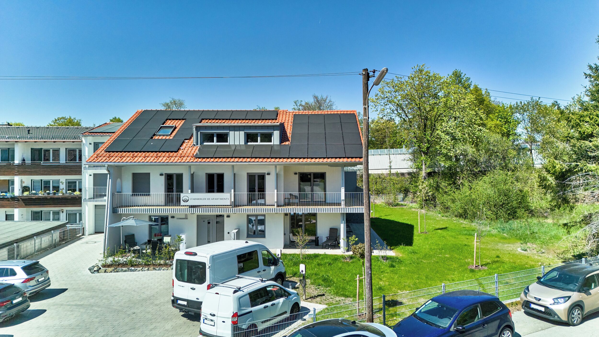 Ein mehrstöckiges Wohngebäude mit einem Ziegeldach, Balkonen und einem Parkplatz mit Autos und einem Wohnmobil.