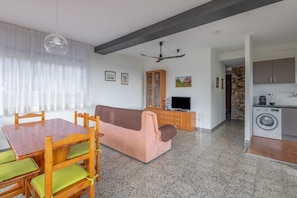 Ein modernes Wohnzimmer mit einer Couch, Esstisch, Stühlen, einem Fernseher und einer Waschmaschine im Hintergrund.