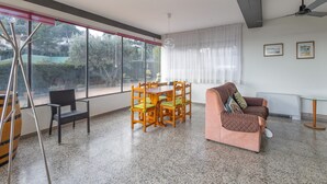 A spacious room with a dining table, chairs, a sofa, and large windows offering a view of the outdoors.