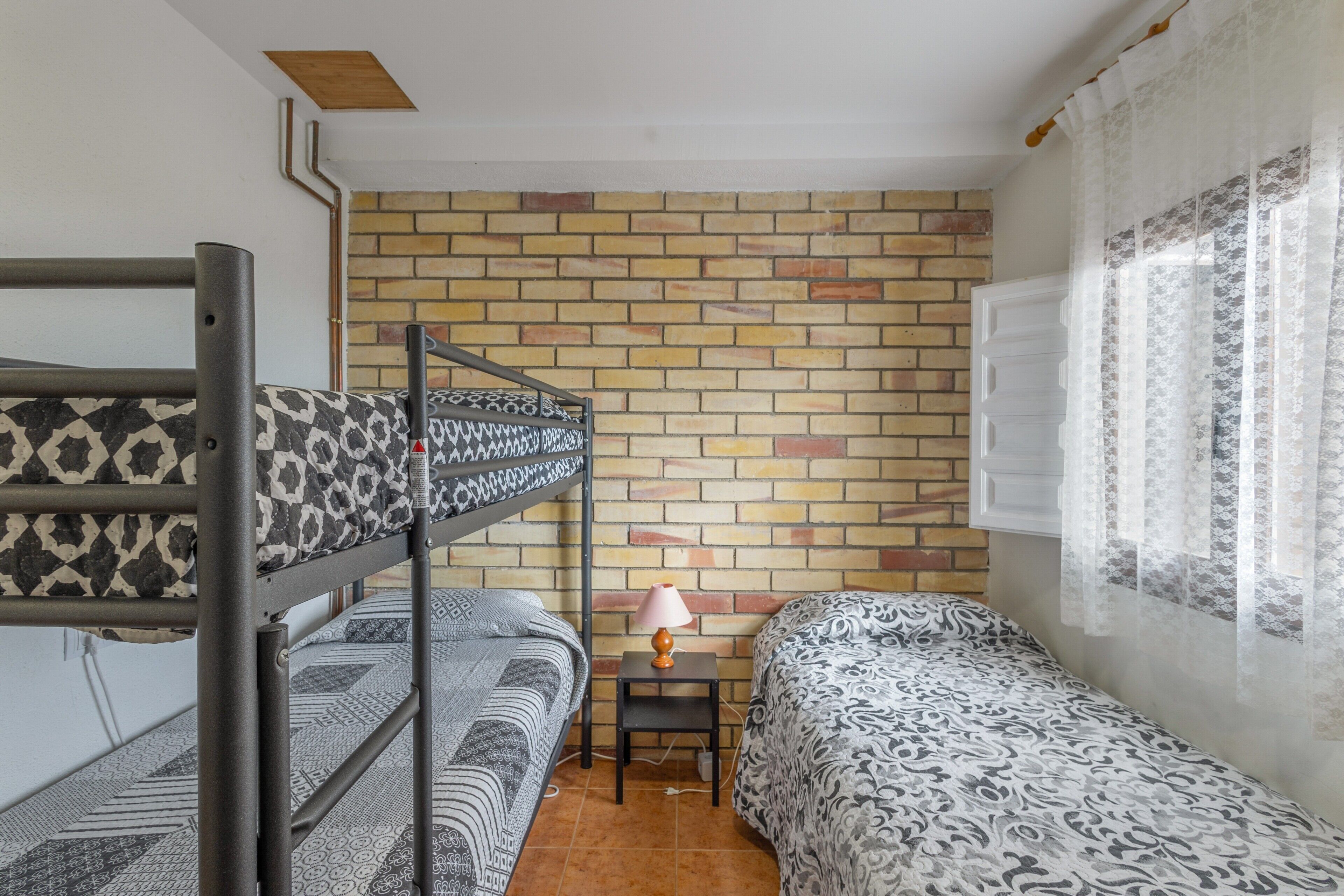 A dormitory room with bunk beds, a small table with a lamp, a brick-patterned wall, and a window with sheer curtains.