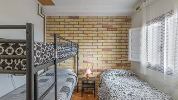 Una habitació de dormitori amb lliteres, una taula petita amb una làmpada, un mur amb relleu de rajola i una finestra amb cortines translúcides.
