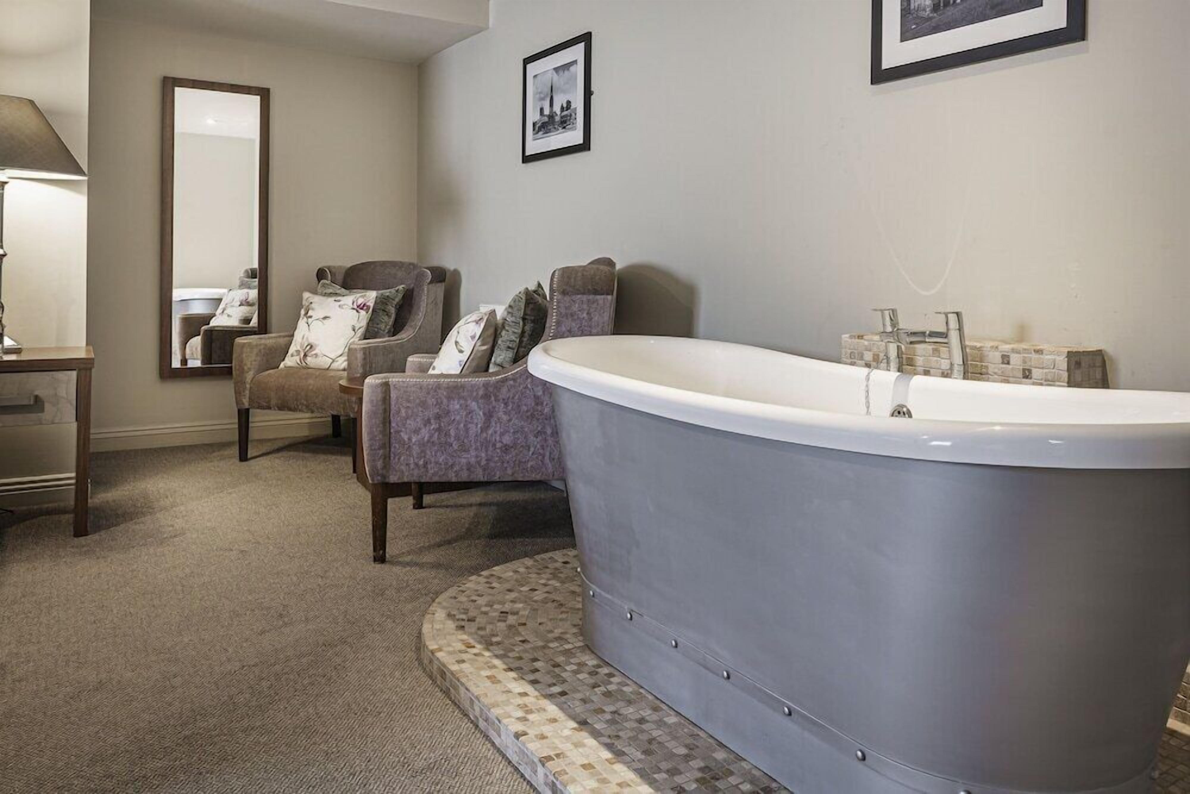 A bathroom with a freestanding bathtub, a chair with cushions, a mirror, and a framed picture on the wall.