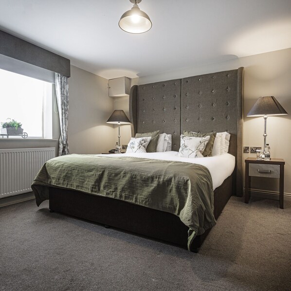 A hotel room with a large bed, a bedside table with a lamp, a window with curtains, and a radiator.