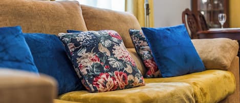 A sofa with floral and solid blue cushions, a wooden coffee table, and a dining area with chairs and a glass.