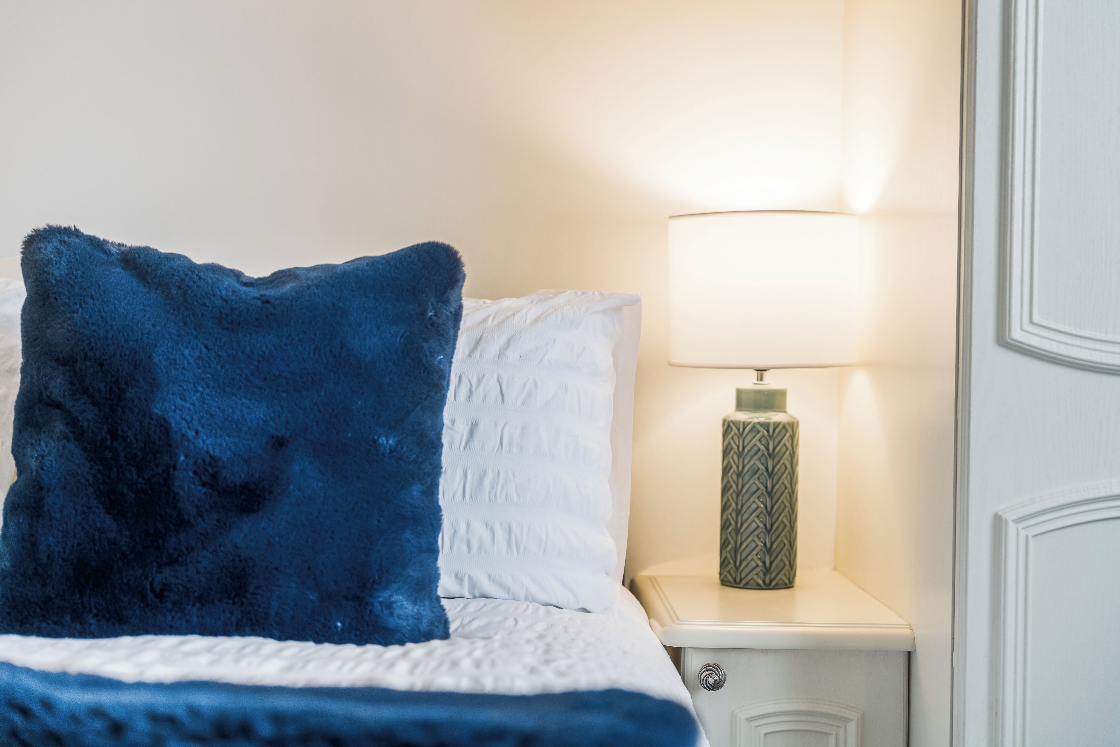 Ein Bett mit einem weiß-blauen Kopfkissen, einer Nachttischlampe und einem weißen Nachttisch.