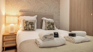A neatly made bed with white linens and two pillows with palm tree designs, a bedside lamp, and a wooden nightstand with a woven basket. - Magnificent Apartment - AC- 2br- 4P - Cannes (Cannes)