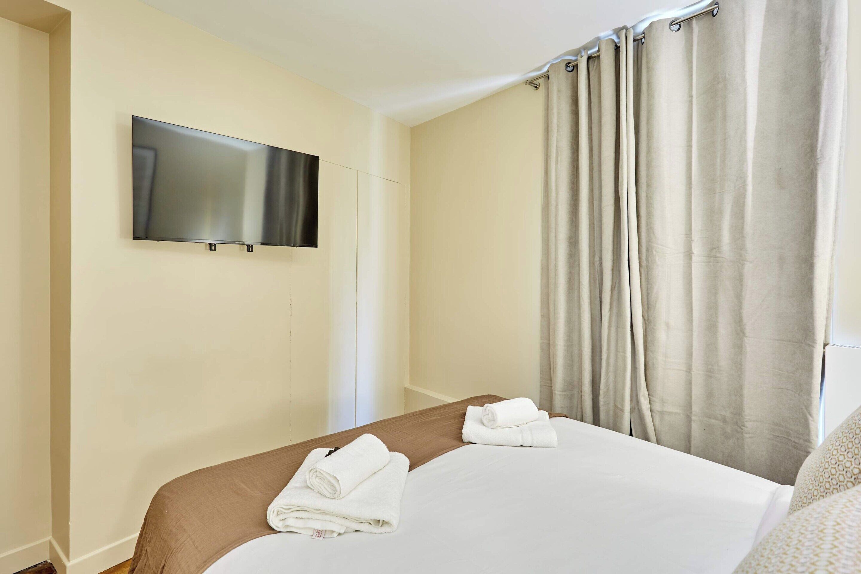A hotel room with a bed, a flat-screen TV mounted on the wall, and a window with curtains.