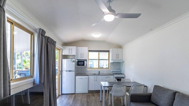 A compact kitchen with white cabinets, a microwave, and a stove. There is a dining area with a table and chairs, and a grey sofa. - Blue Dolphin Holiday Resort (Yamba)