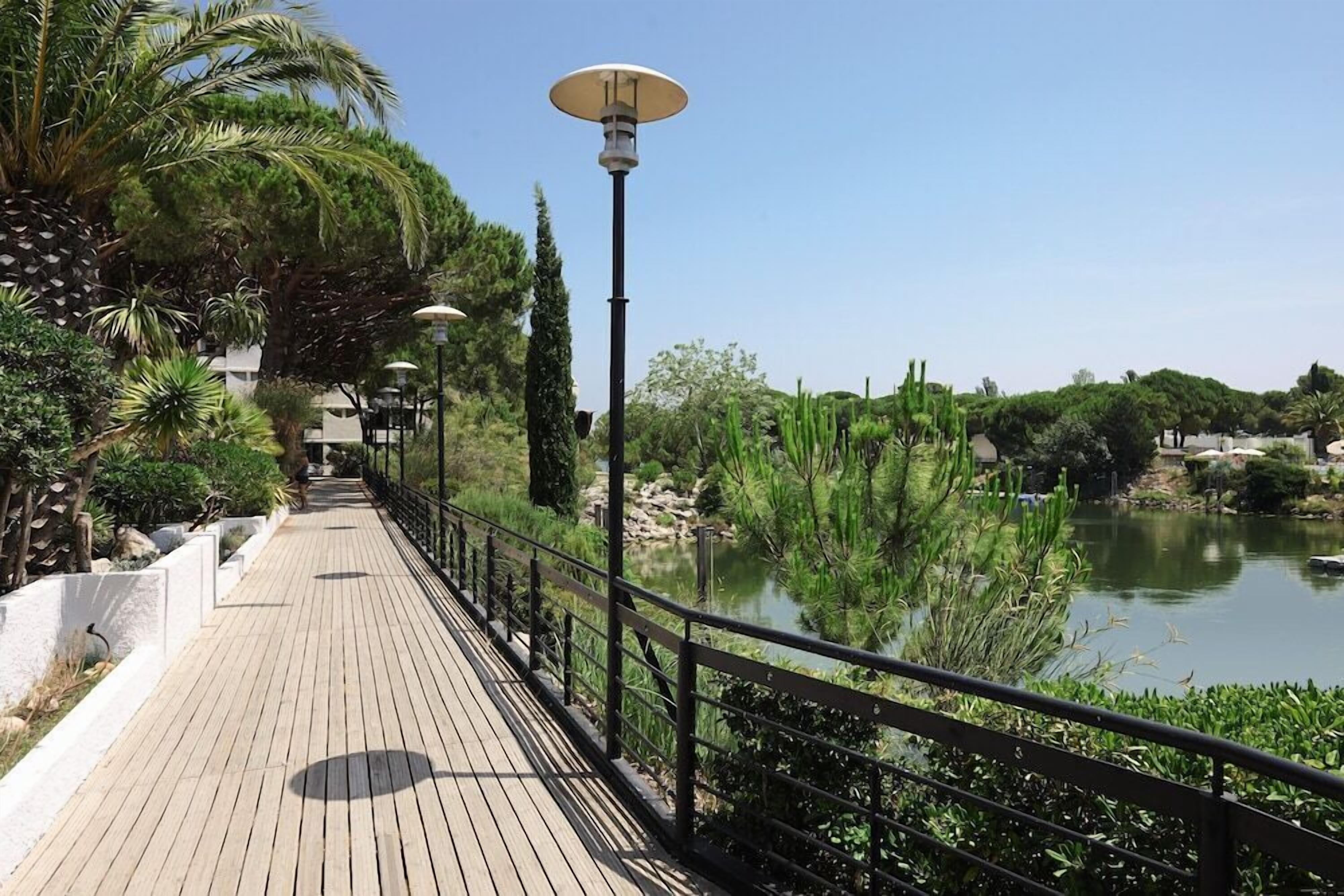 Une promenade en bois dotée d’une rambarde métallique, qui mène à une étendue d’eau bordée de verdure et de cactus.