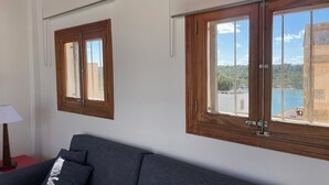 Une pièce avec un canapé gris foncé, deux fenêtres à cadre en bois dotées de poignées en métal et un mur blanc.