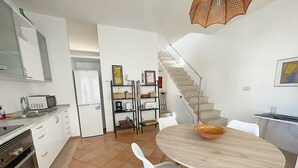 Une cuisine moderne avec des armoires blanches, un four à micro-ondes encastré et un réfrigérateur. On y trouve un coin repas avec une table ronde et des chaises blanches. Un escalier mène à l’étage supérieur.