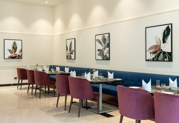 A modern dining area with a long table, purple chairs, and abstract wall art. - Palm Meadows Resort (Bengaluru)