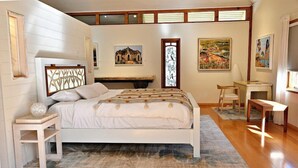 Un dormitorio con cama, mesita de noche, escritorio y silla. Hay cuadros en las paredes y una ventana con vistas.