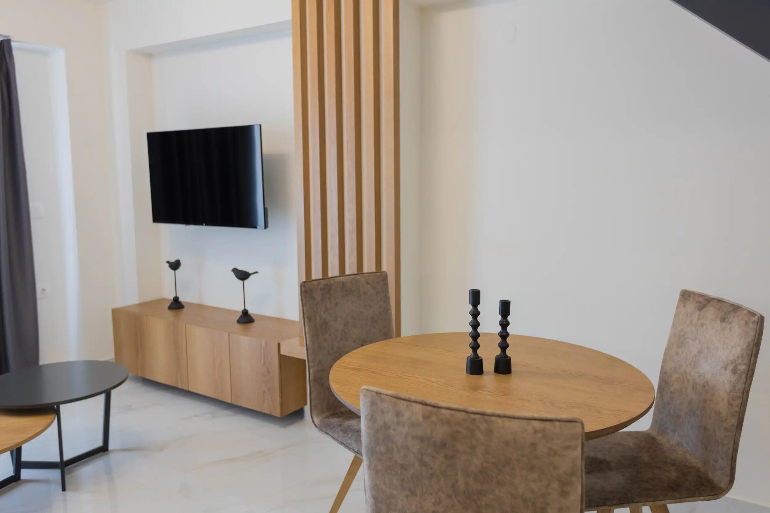 Ein modernes Wohnzimmer mit einem runden Holztisch, zwei gepolsterten Stühlen, einem Flachbildfernseher an der Wand und einer Holzkommode mit zwei lampenförmigen Vogel-Leuchten.