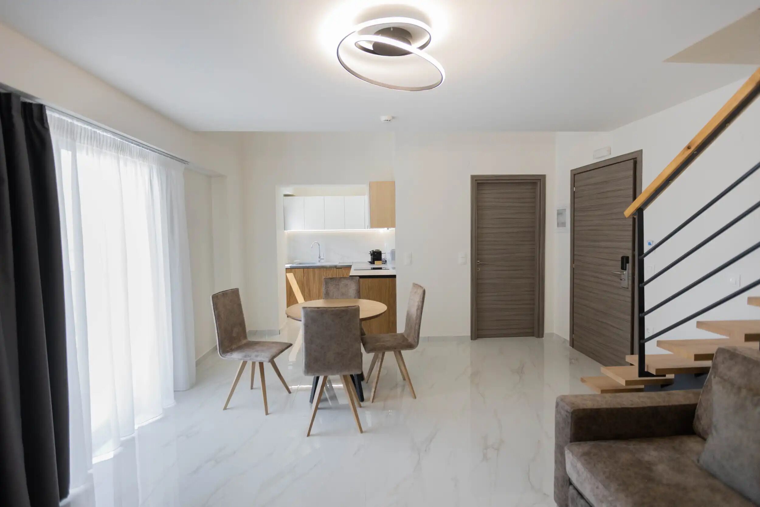 A modern living room with a dining area, a staircase, and a kitchen in the background.