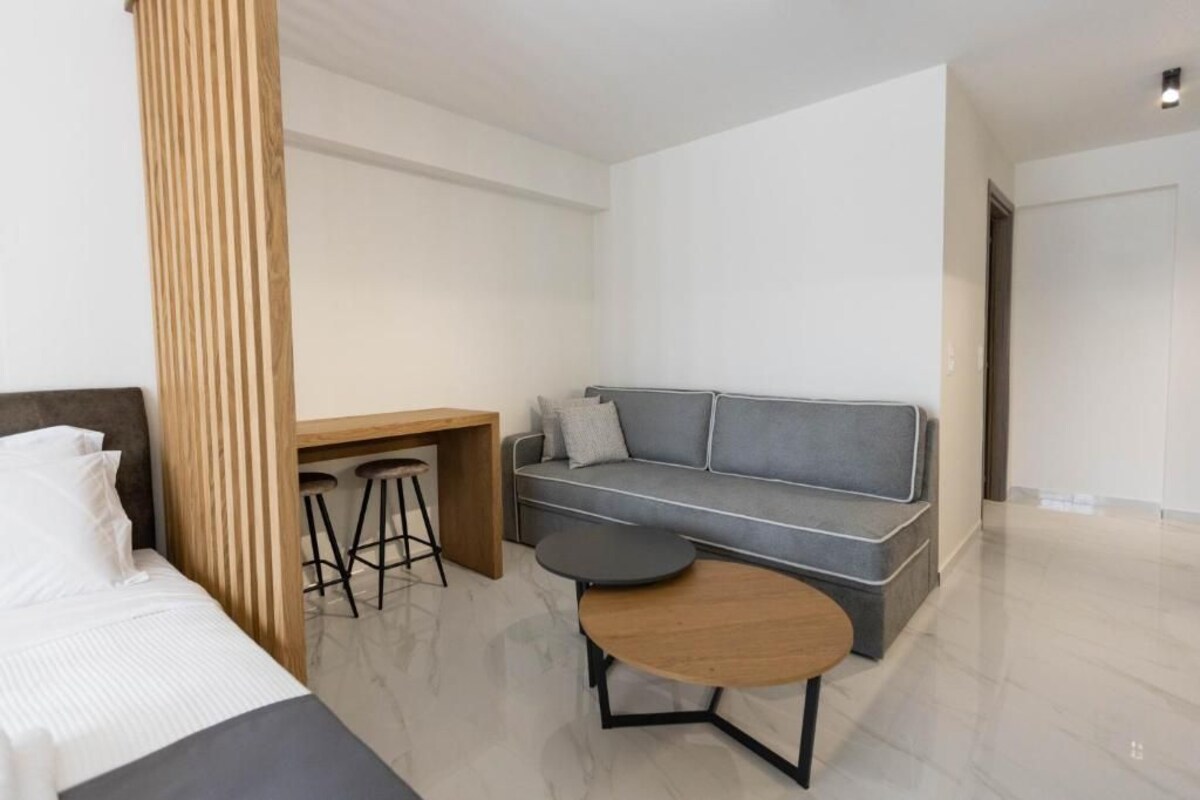 A modern hotel room with a bed, a grey sofa, a wooden desk, two stools, and a small round table.