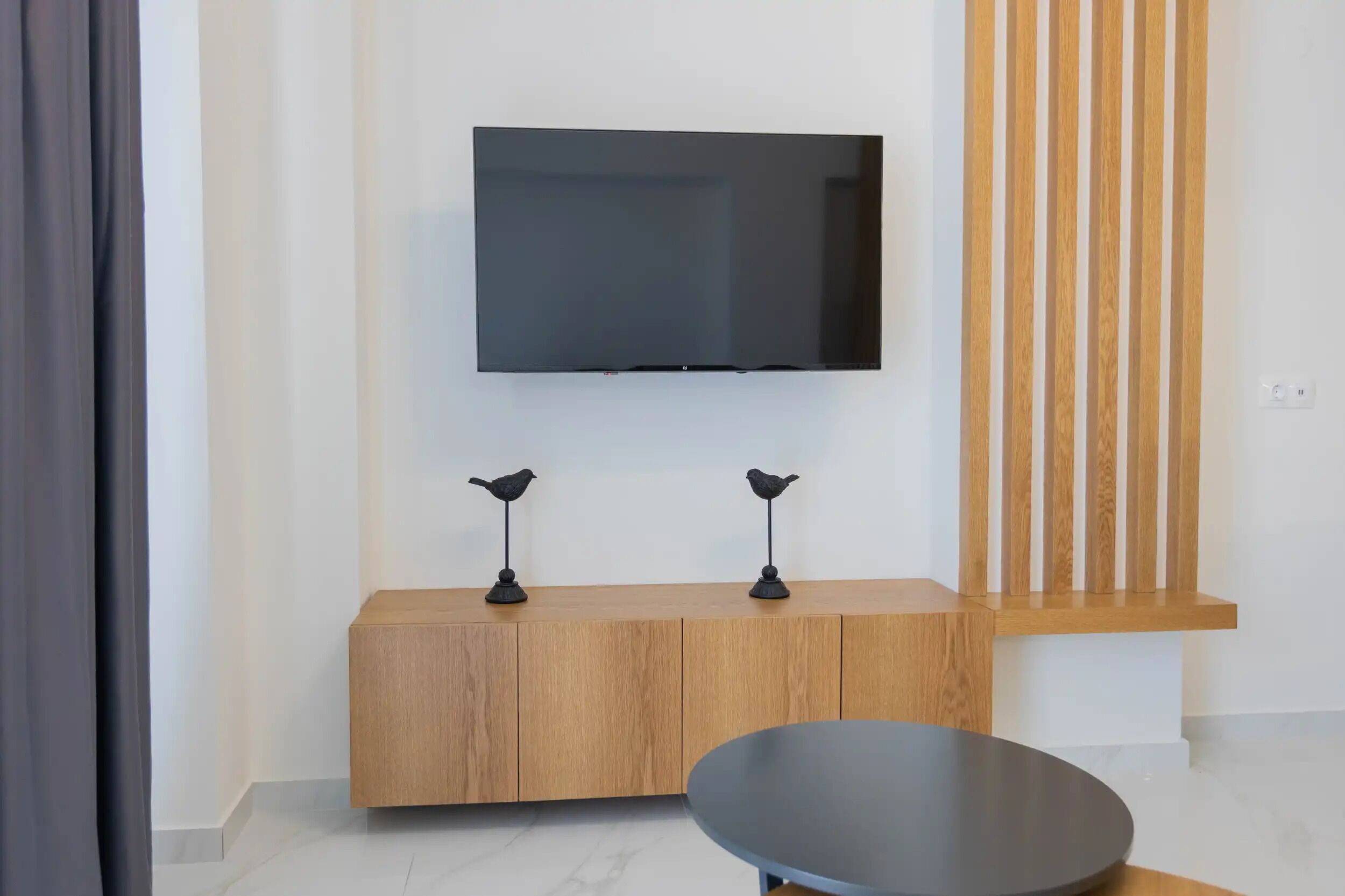 A modern living room with a flat-screen TV mounted on the wall, a wooden console table with two bird-shaped lamps, and a round coffee table.