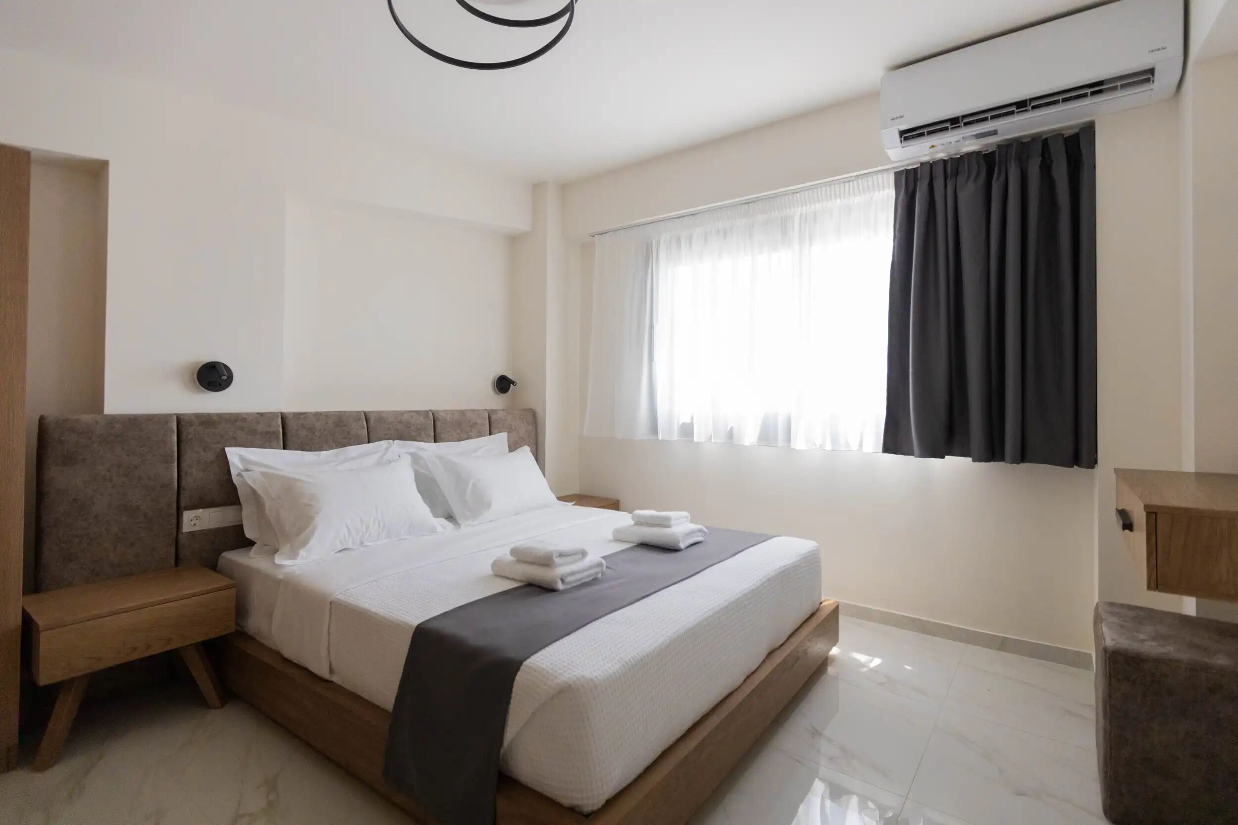 A modern hotel room with a large bed, a bedside table, an air conditioning unit, and a window with curtains.
