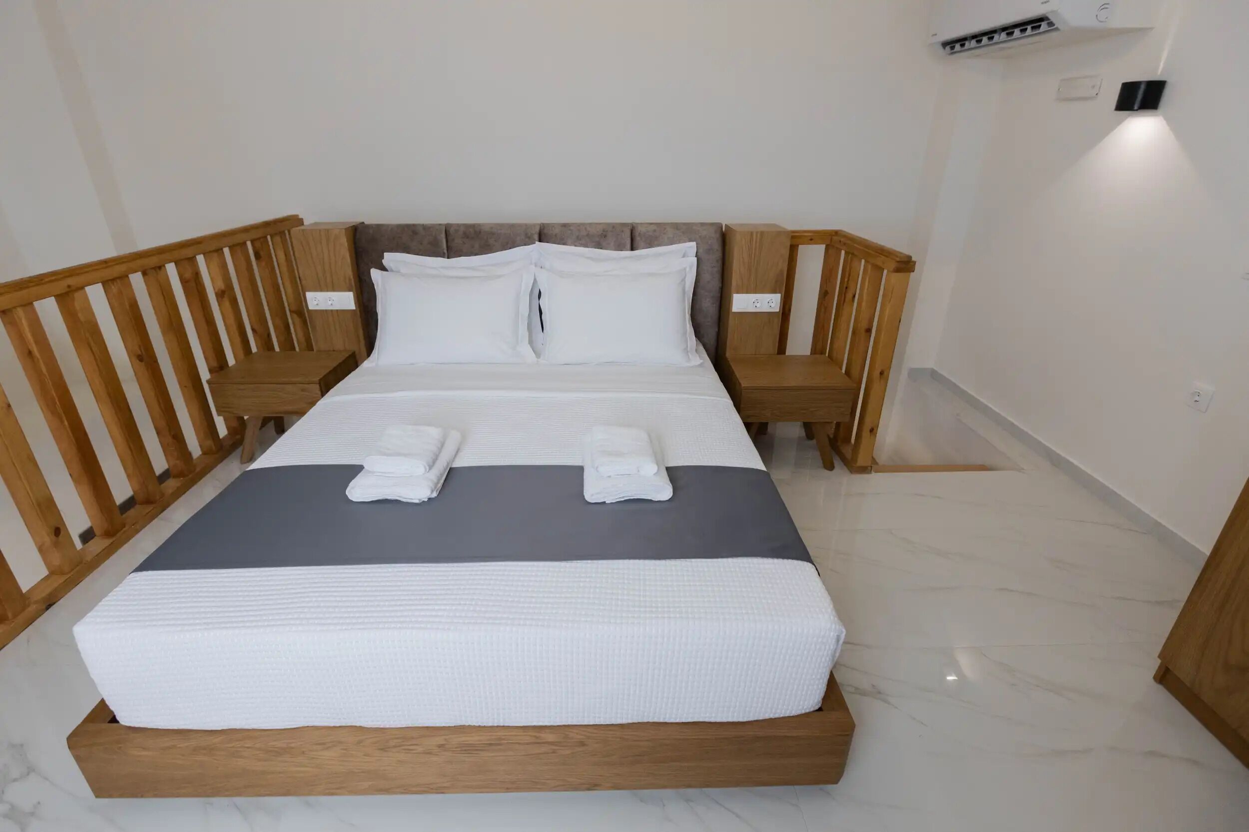 A neatly made bed with white linens and two folded towels, a wooden headboard, and a marble floor.