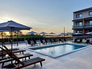 A rooftop pool area with sun loungers and umbrellas, a swimming pool, and a view of a modern building.