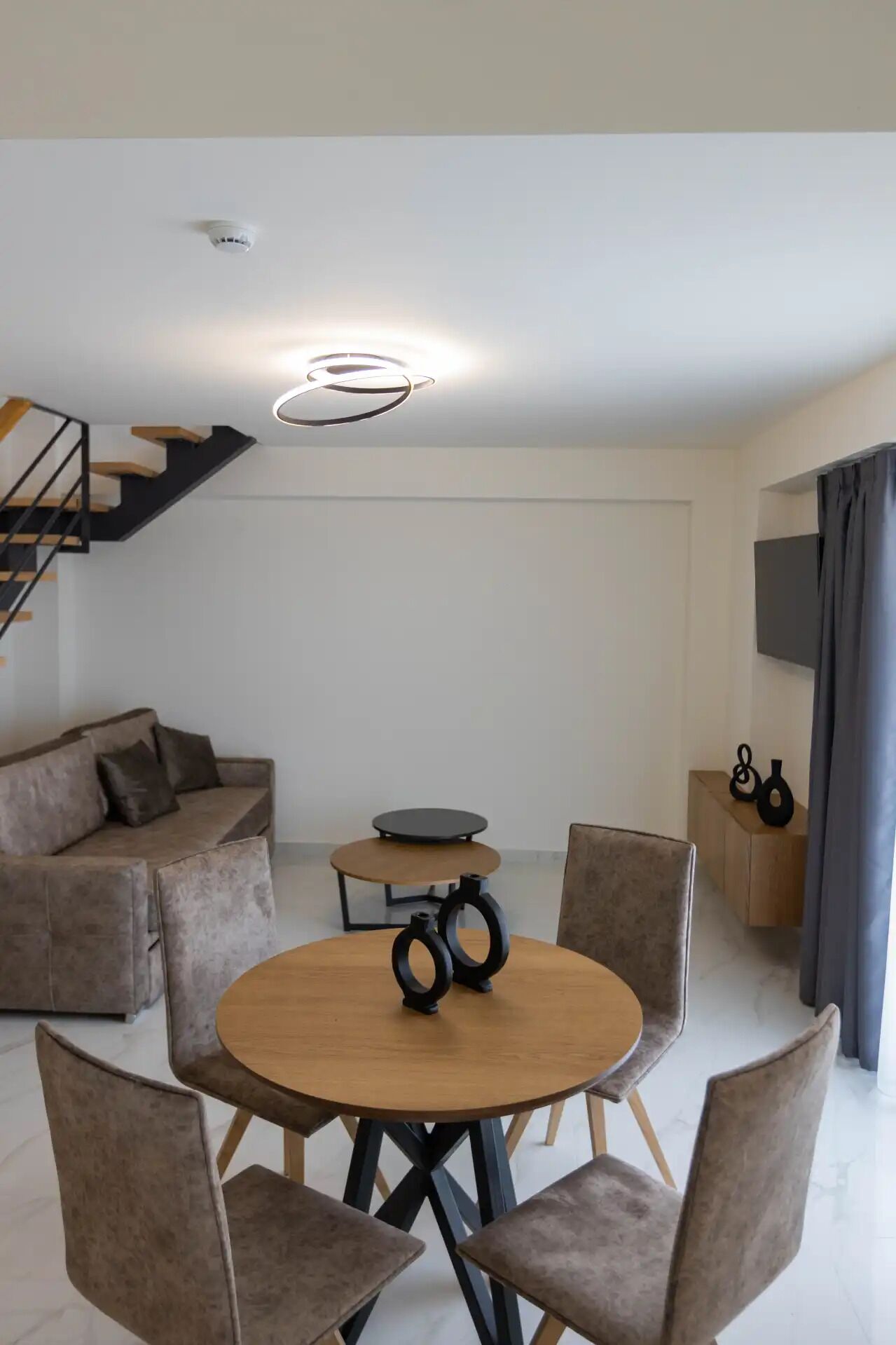 Una sala de estar moderna con una mesa redonda de madera, sillas con cojines, un sofá, una pequeña mesa con superficie negra, una escalera y una lámpara de techo.