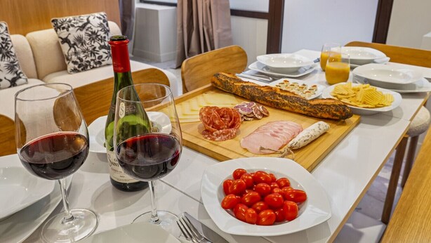 Ein Esstisch, gedeckt mit Weingläsern, einer Weinflasche, verschiedenen Käsesorten, Wurstwaren, einem Baguette, Cocktailtomaten und einer Portion Pommes.