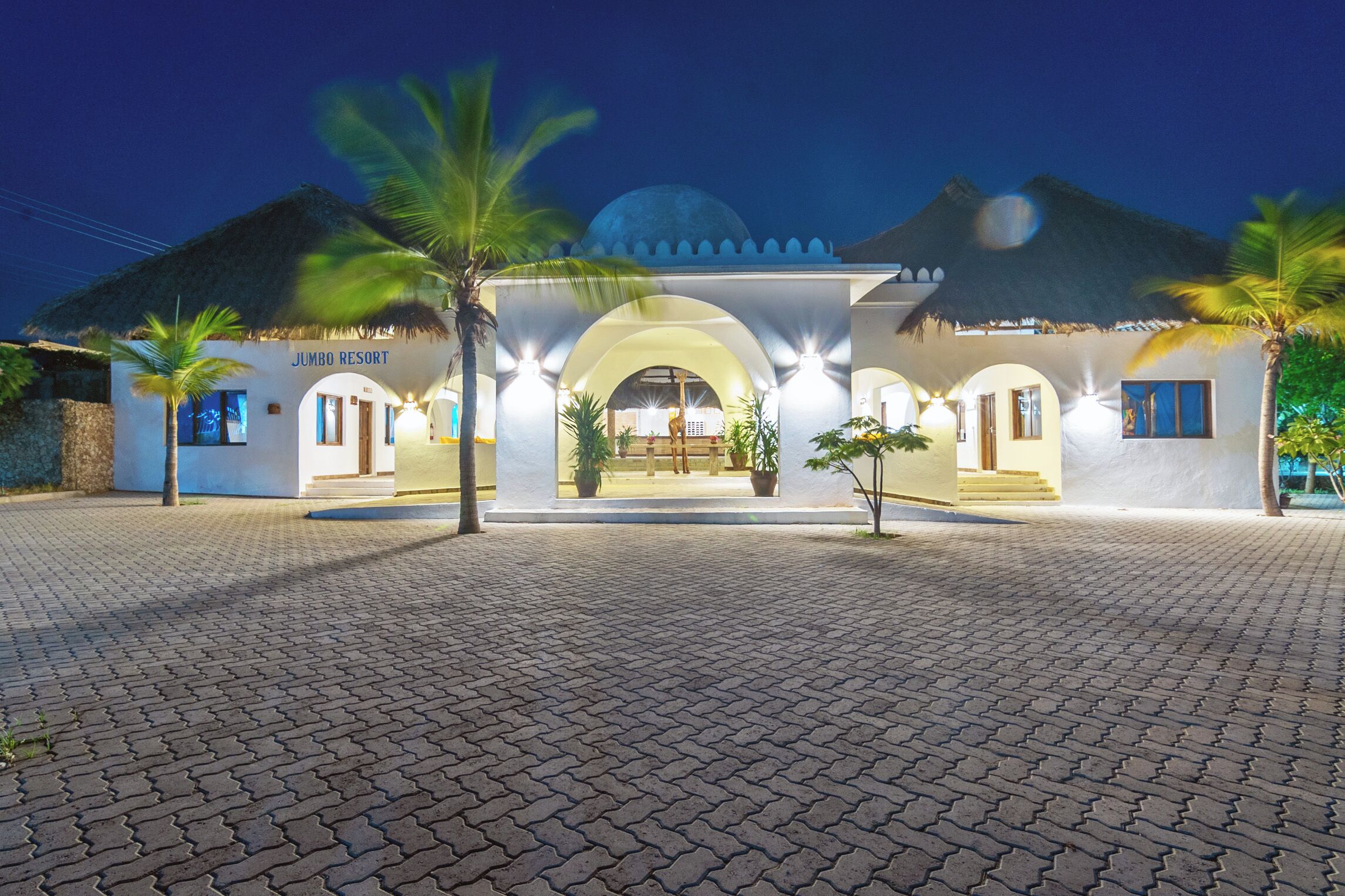 A resort with a thatched-roof entrance, palm trees, and a paved walkway.