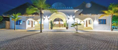A resort with a thatched-roof entrance, palm trees, and a paved walkway.