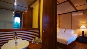 Hotellihuone, jossa on puinen sänky, yöpöytä lampun kanssa, suuri peili sekä kylpyhuone, jossa on pesuallas ja hygieniatarvikkeita.