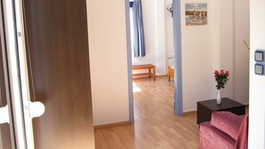 Un pasillo con suelo de madera, una puerta con marco azul, un banco, una pequeña mesa con un florero y un cuadro en la pared.