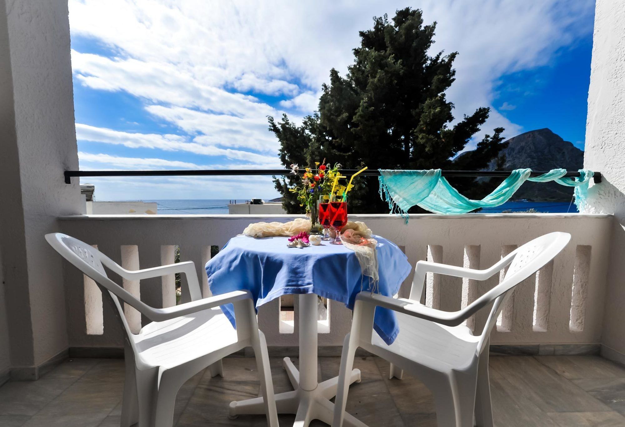 A balcony with a table covered in a blue cloth, white plastic chairs, and a vase with flowers.