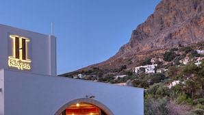 Un hotel con un llamativo letrero en forma de 'H' y 'Kalypsos' escrito debajo, frente a un paisaje montañoso.