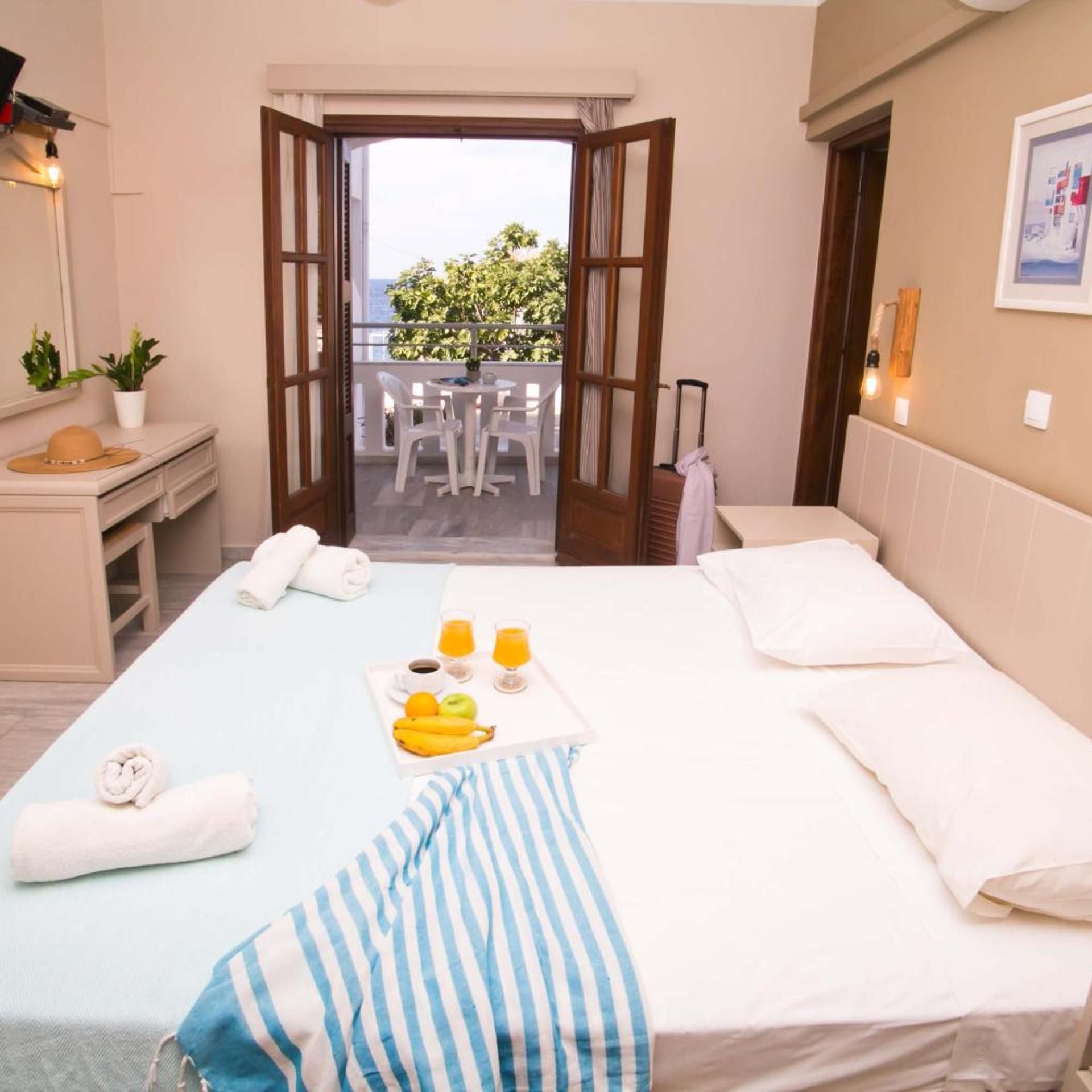 A hotel room with a bed, a small desk, a balcony with chairs, and a view of greenery.