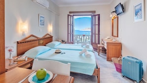 Habitación de hotel con dos camas, un escritorio, una silla, una mesa con una fuente de fruta, una maleta y vista al mar.