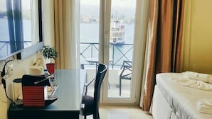 Ett hotellrum med en säng, ett skrivbord med en röd apparat på, en stol, ett litet bord och utsikt över en vattenyta genom en glasdörr.