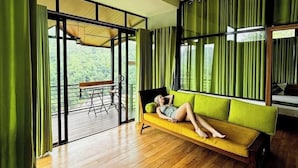 A room with a wooden floor, a yellow sofa, and a view of greenery through large windows. - Aqua Dunhinda Villa & Nature Retreat (Gampola)