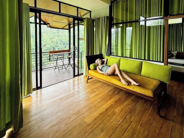 A room with a wooden floor, a yellow sofa, and a view of greenery through large windows. - Aqua Dunhinda Villa & Nature Retreat (Gampola)
