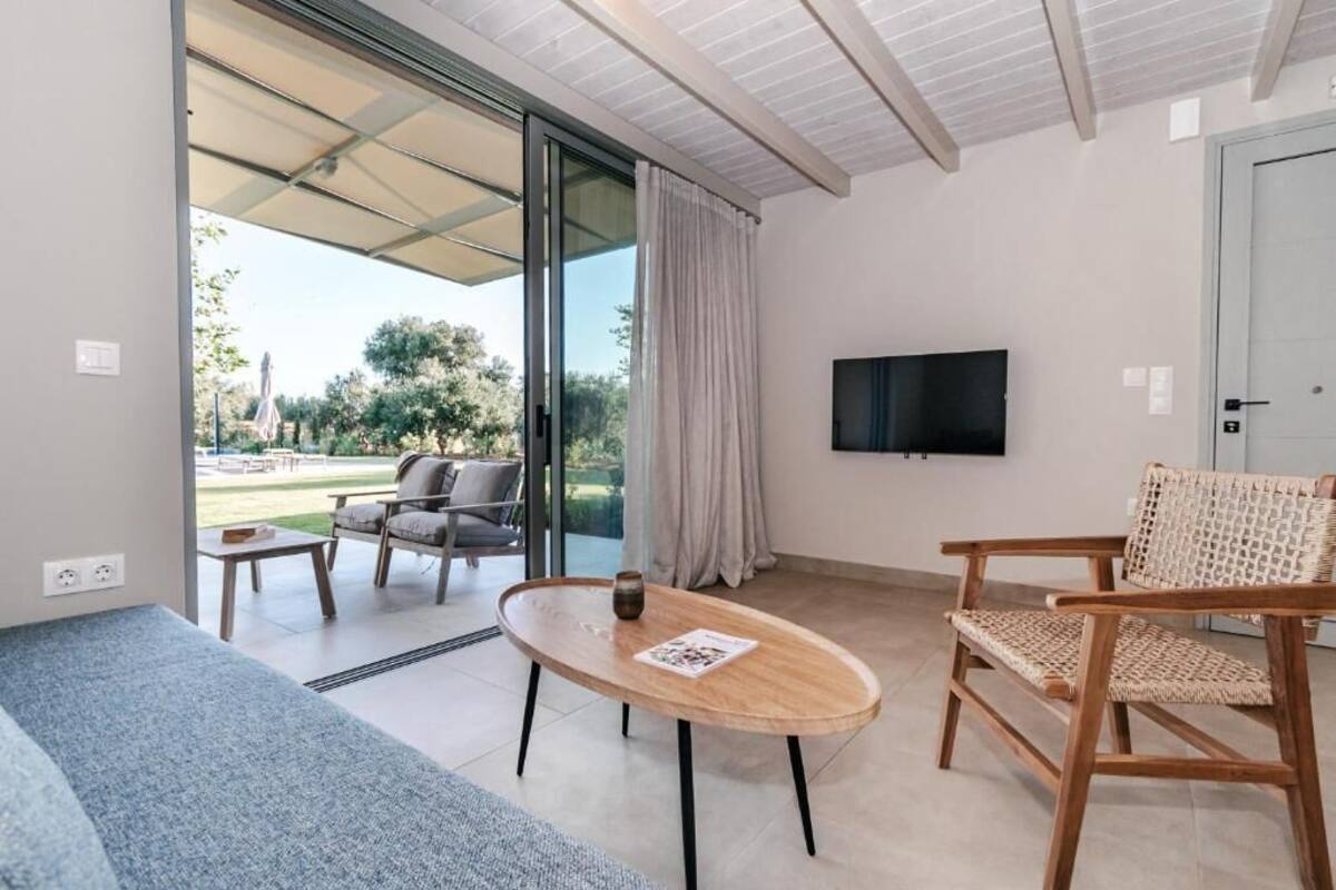 Una sala moderna con un sofá, una mesa de madera y una silla de mimbre. Hay un televisor montado en la pared y un ventanal corredero que da a un espacio exterior.