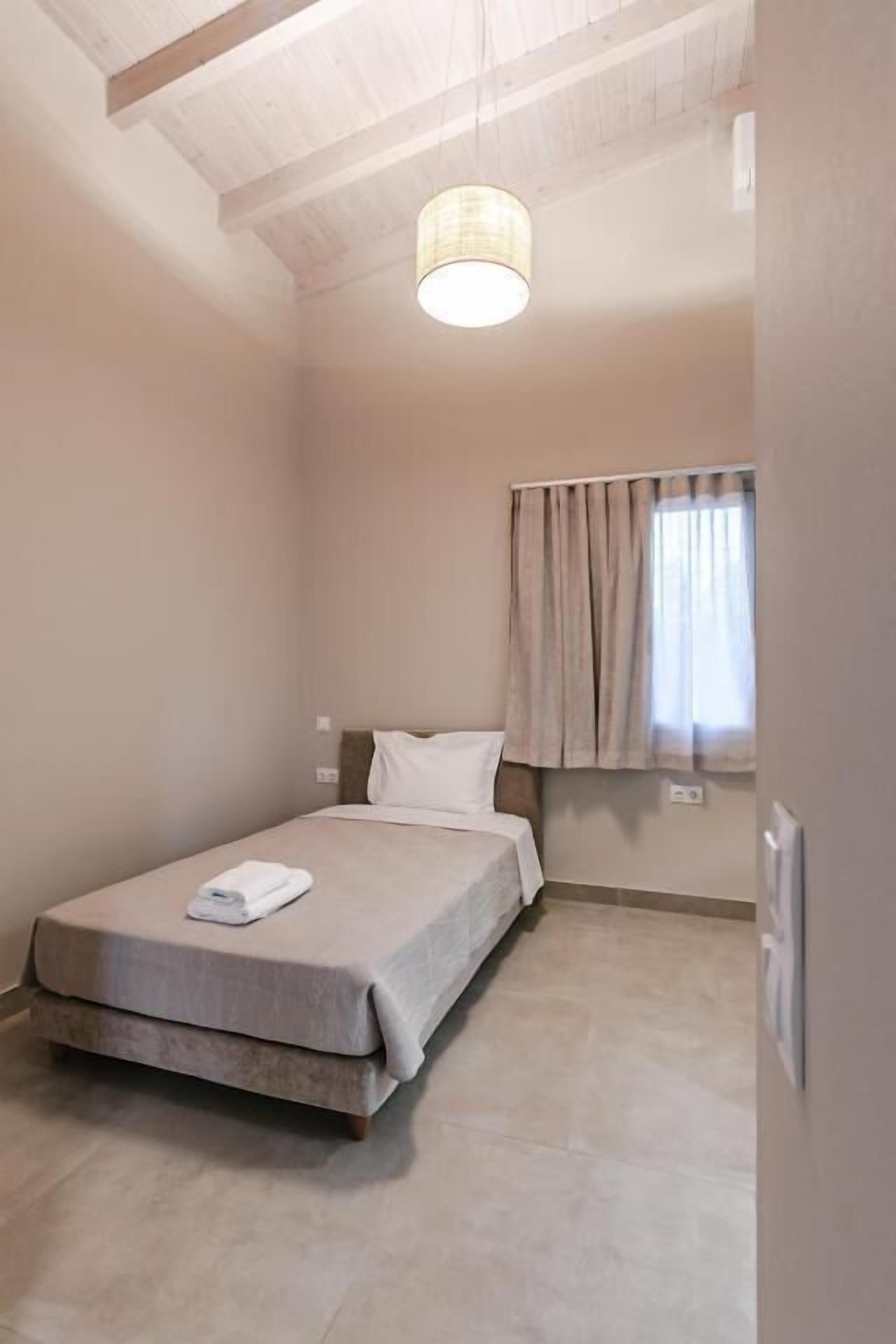 Una camera piccola con un letto singolo, arredamento dai toni neutri, una ventola a soffitto e una finestra con tende.