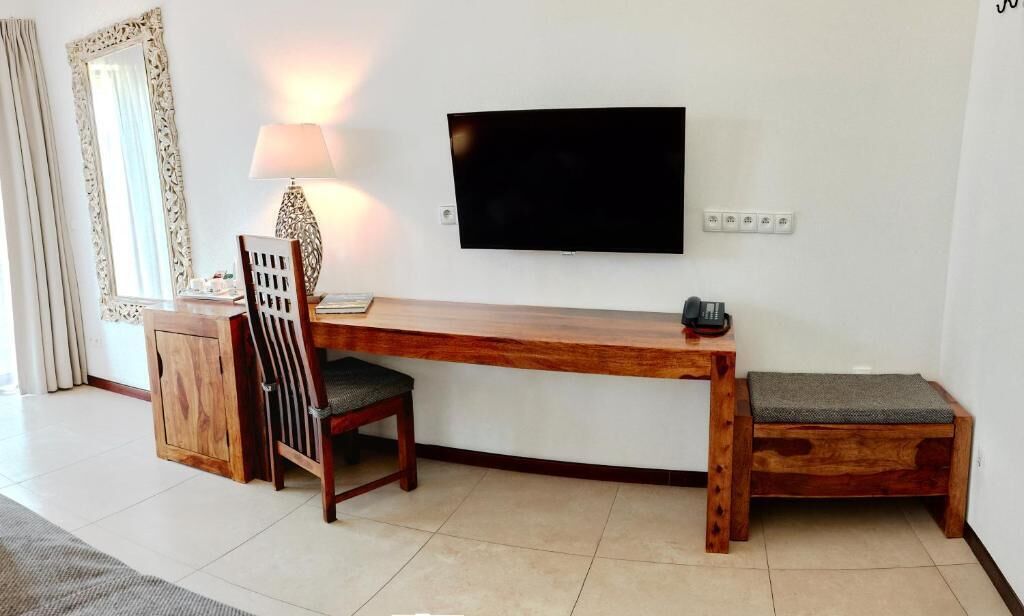 A wooden desk with a chair and a bench, a television mounted on the wall, and a lamp on the desk.