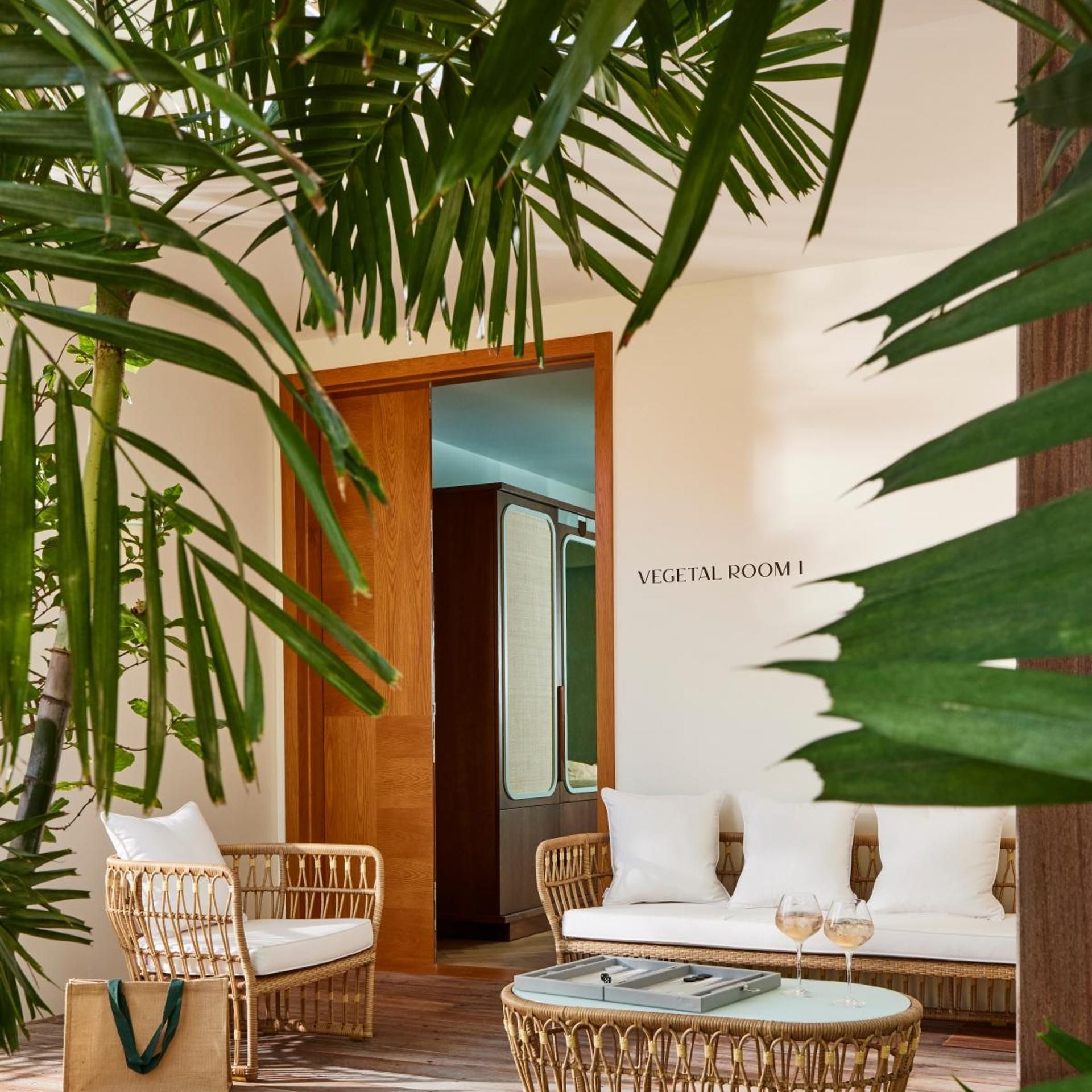 A room with a wicker sofa, a wicker coffee table, and a wicker chair. There is a door leading to a room labeled 'VEGETAL ROOM 1'.