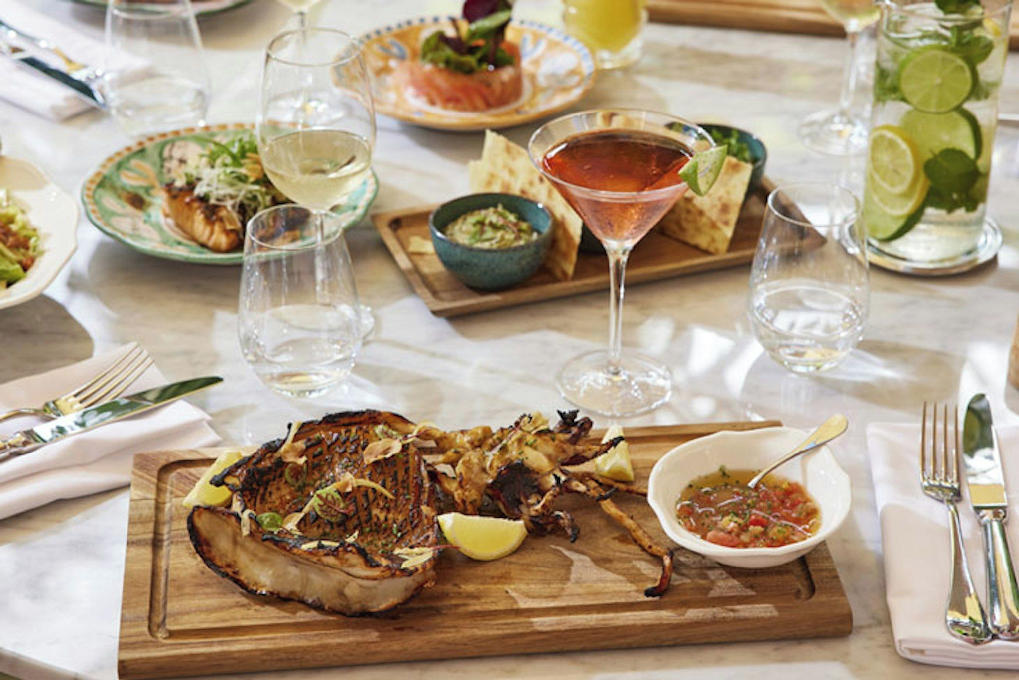 Una mesa de comedor con diversos platillos, incluyendo carne a la parrilla, un martini y un tazón de salsa.
