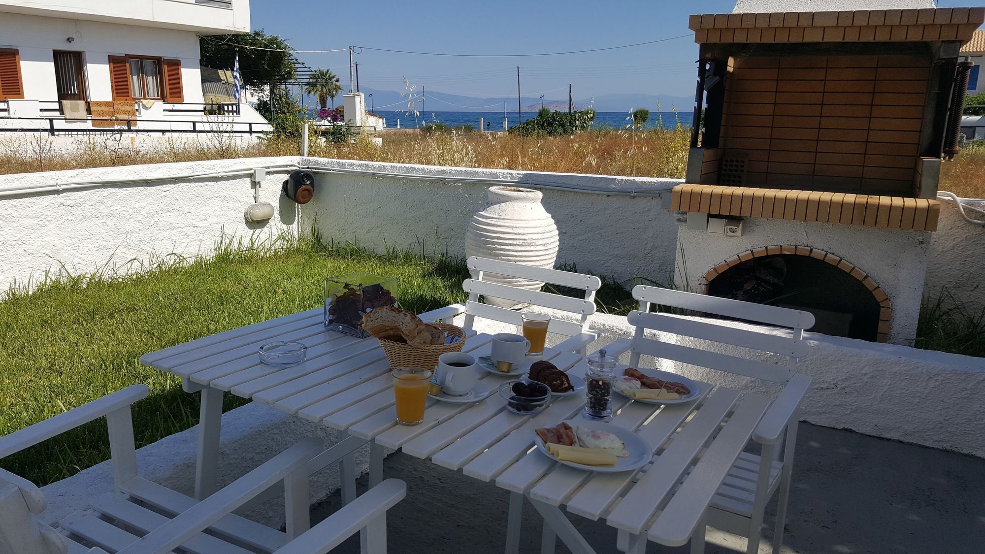 A table set with breakfast items, a white bench, and a brick oven, with a view of the sea and mountains in the background.