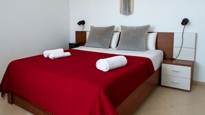 Un letto rifatto con cura, con una coperta rossa, due cuscini e due asciugamani arrotolati. È presente un comodino con cassetti e una lampada fissata alla parete.